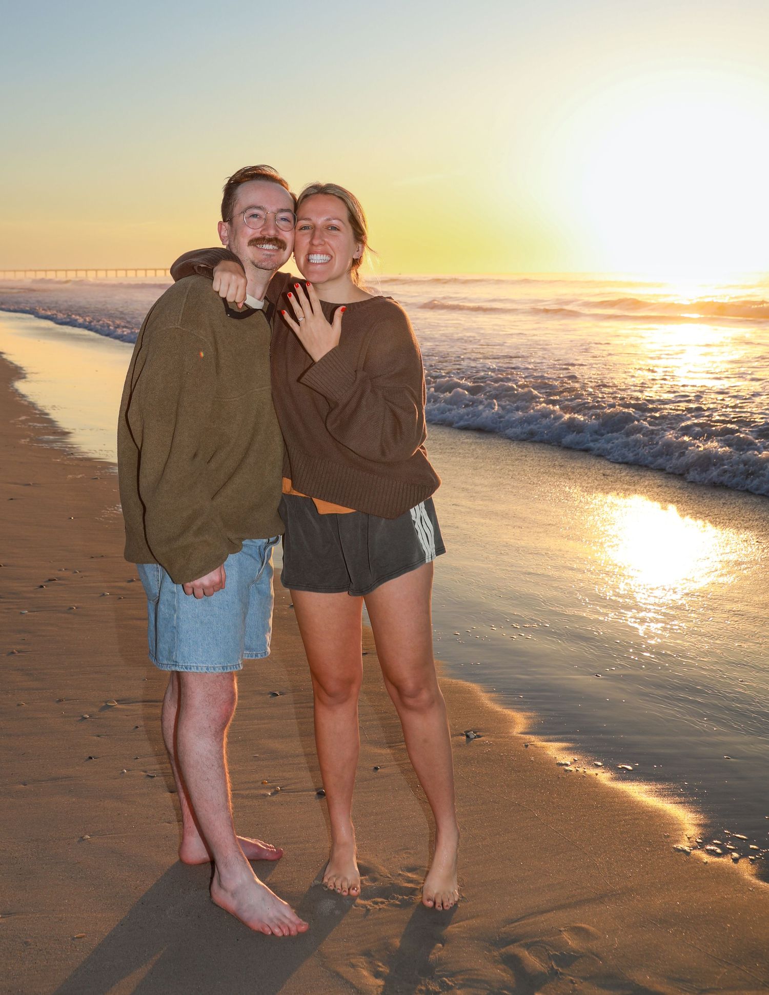 Couple smiling together on a sandy beach at sunset, waves rolling in behind them, woman showing off engagement ring.