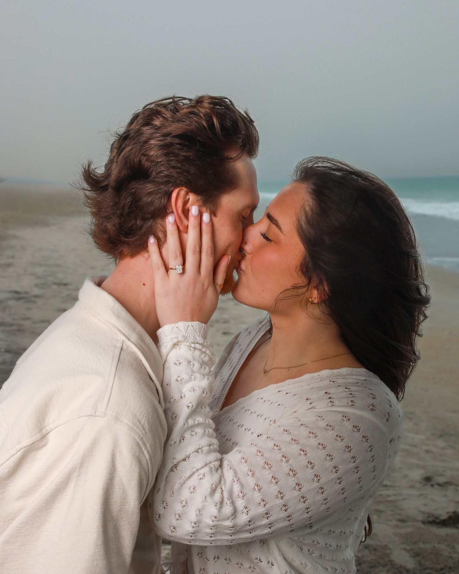 Couple sharing a romantic kiss on a foggy beach, woman wearing an engagement ring and white lace dress.