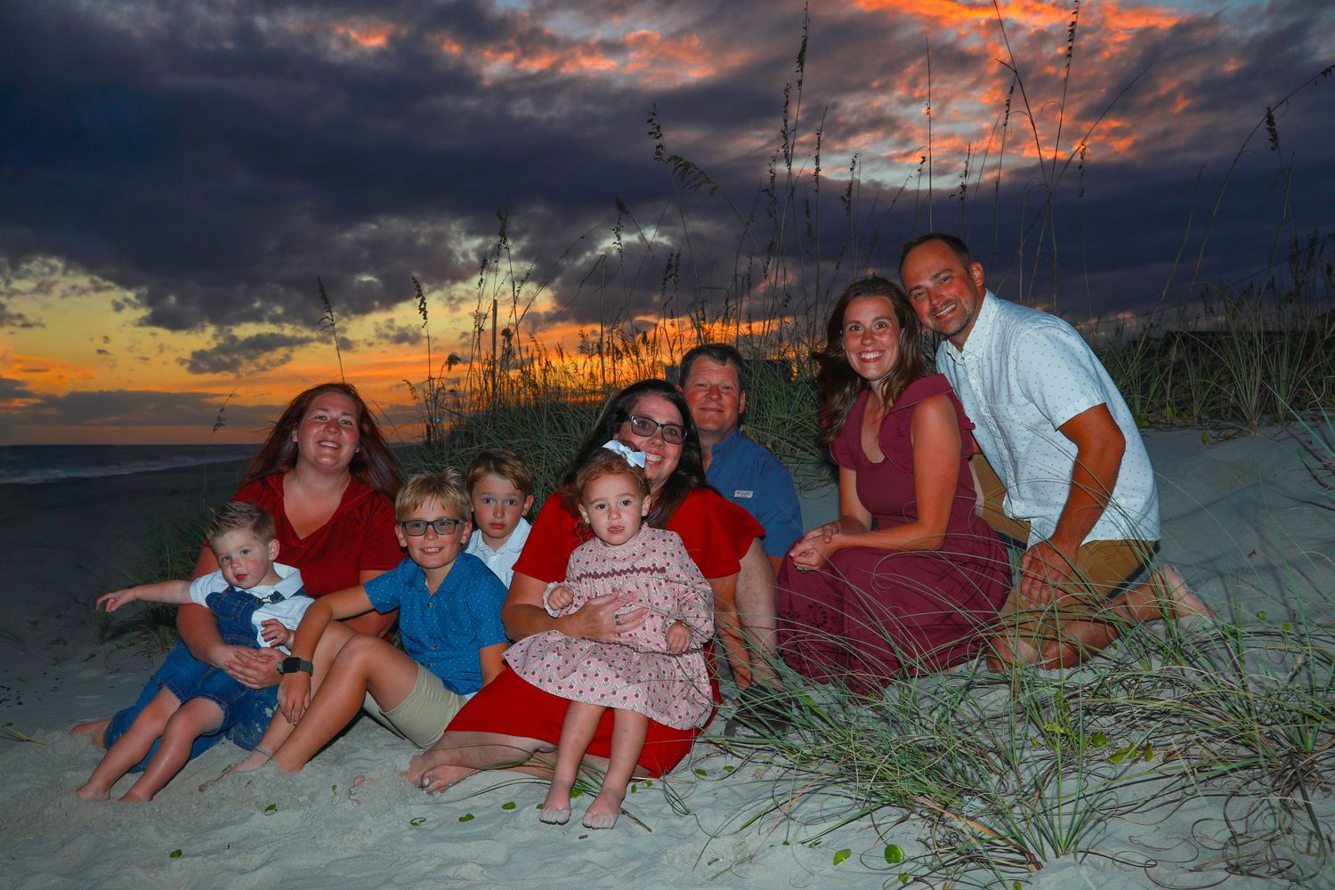 Family portrait at sunset on a sandy beach with dramatic colorful sky, featuring two adults and several children sitting together.