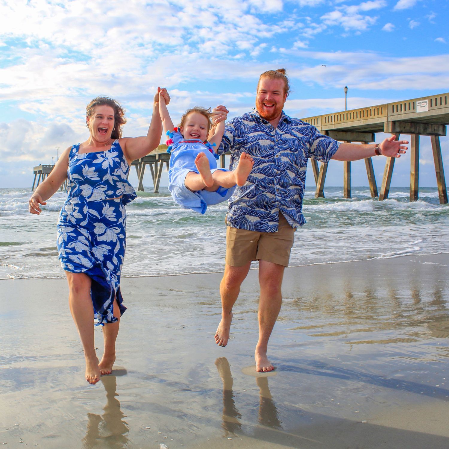 Family playing and splashing in ocean waves near a pier wearing matching blue outfits.
