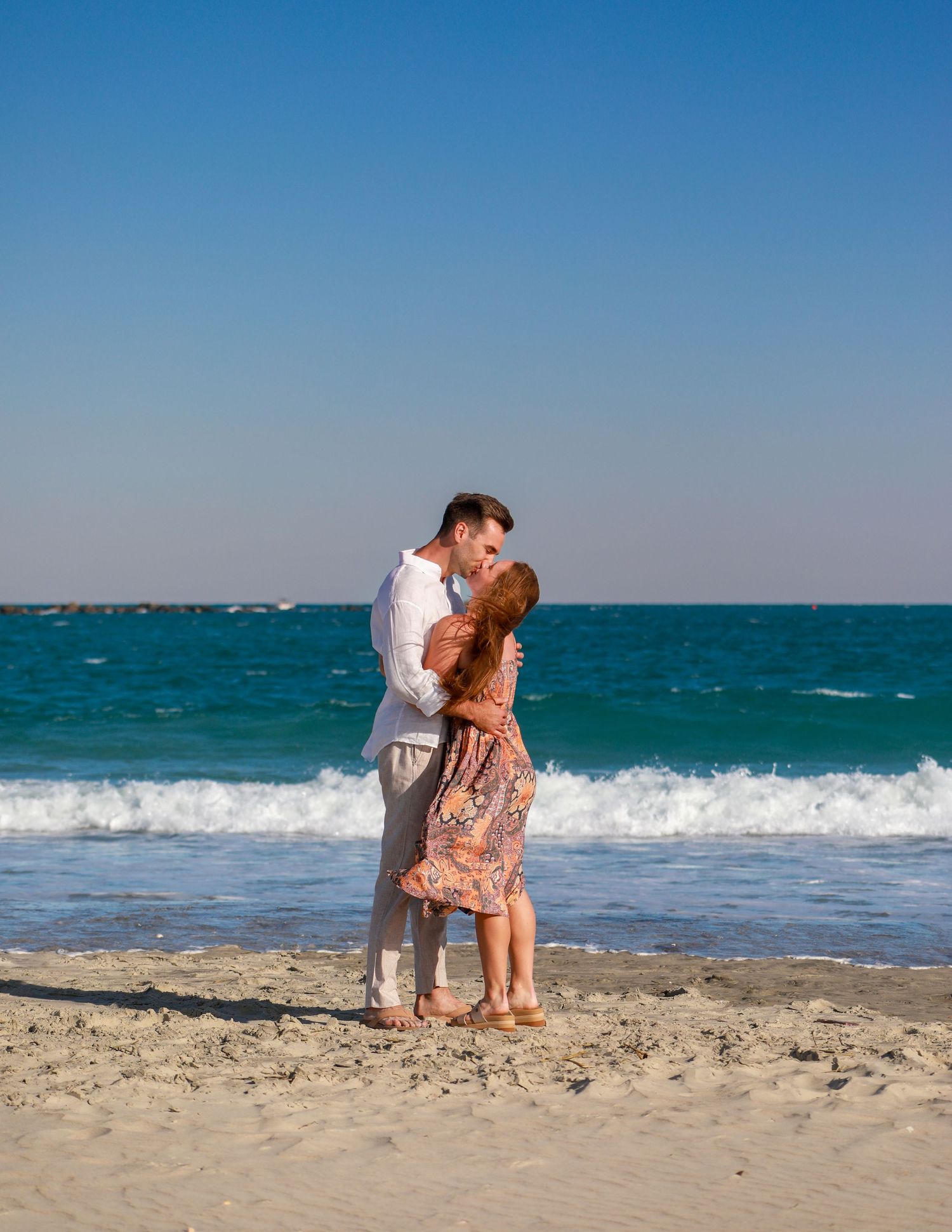 Couple kissing on a sandy beach with turquoise ocean waves and clear blue sky backdrop.