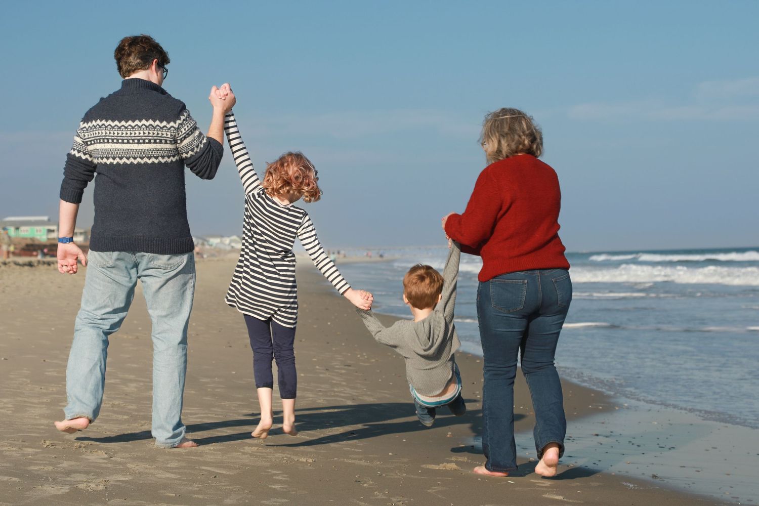 Family of four walking barefoot on a sandy beach, parents swinging young child between them near ocean waves.