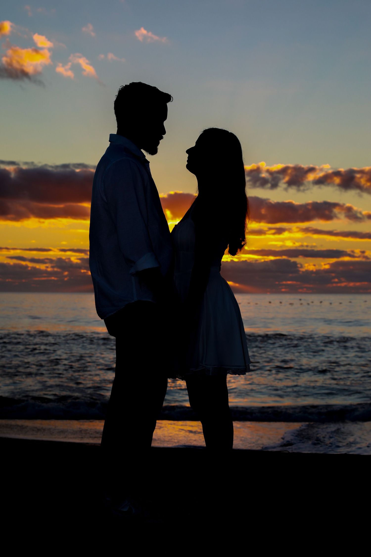 Romantic silhouette of a couple facing each other on a beach at sunset with colorful clouds over the ocean.