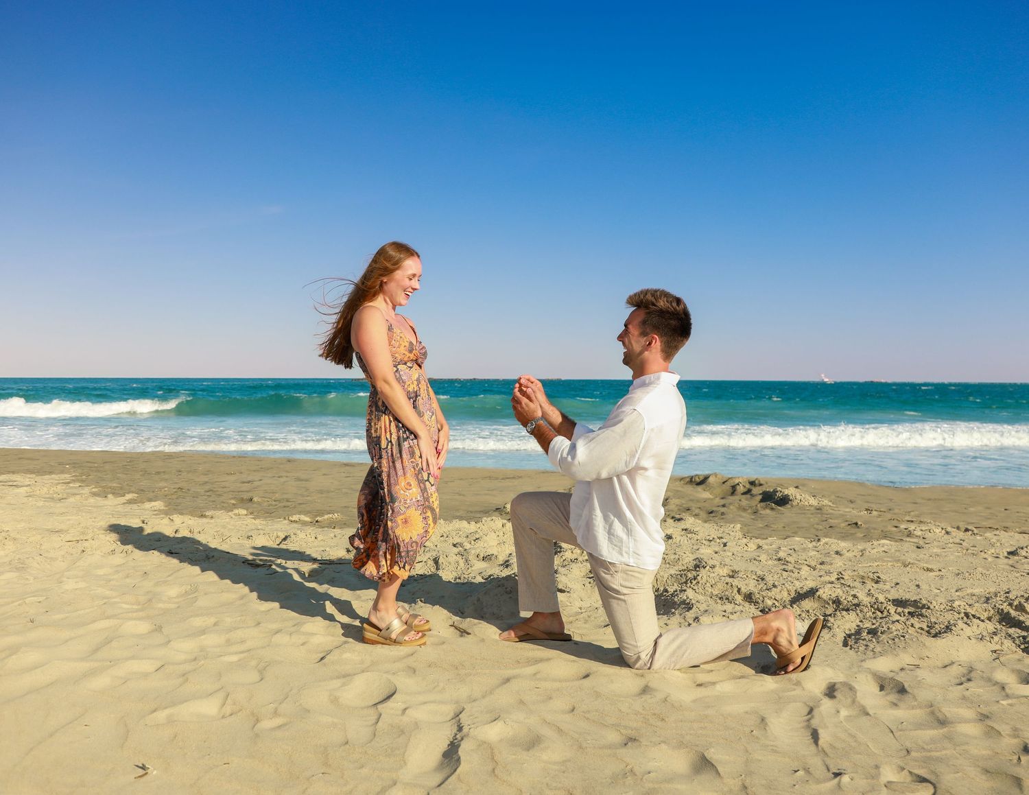 Man proposing to woman on a sandy beach with turquoise ocean waves in the background on a sunny day.