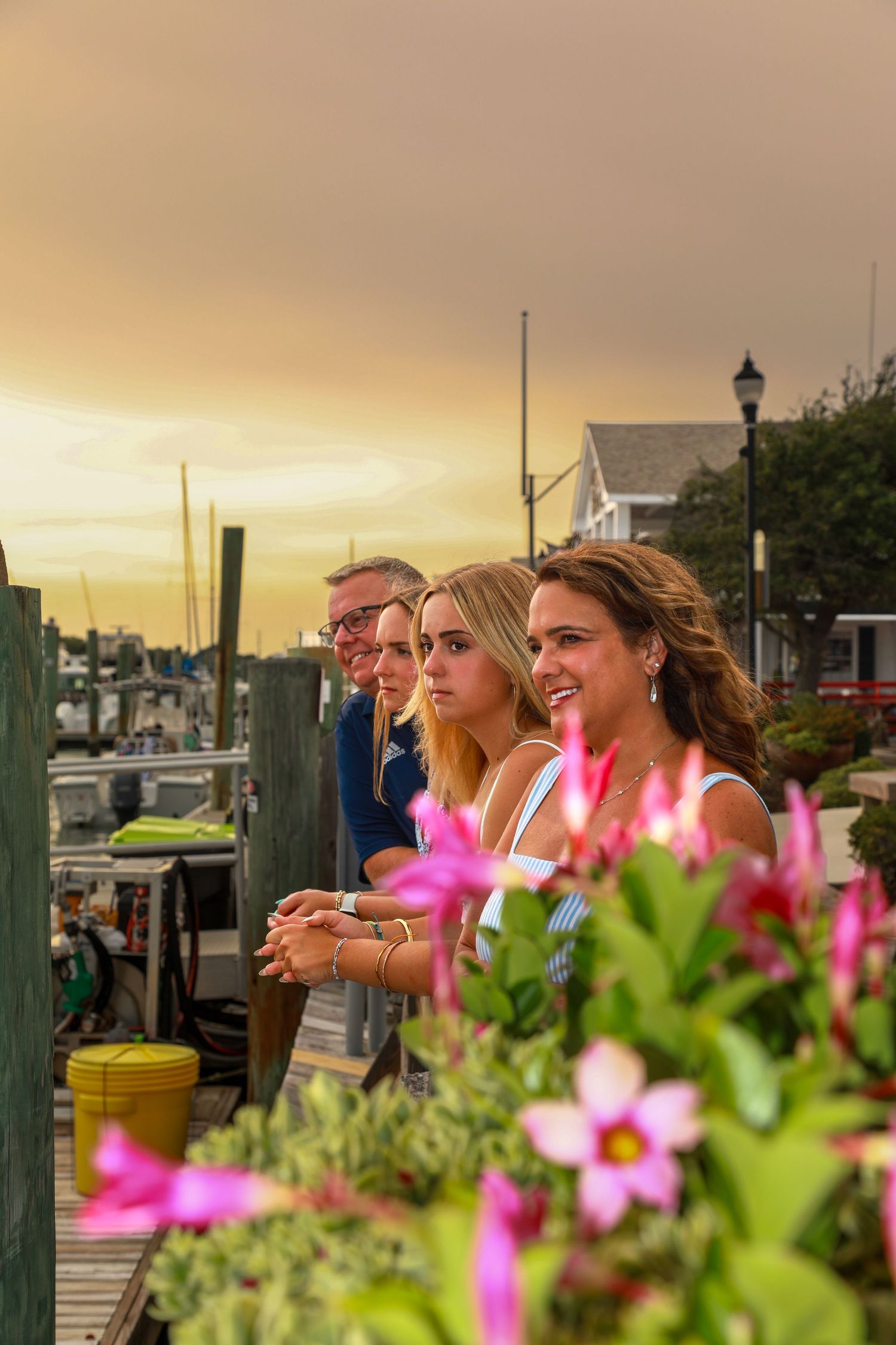 Family enjoying a stunning waterfront sunset from a dock, with pink flowers in the foreground at a marina.