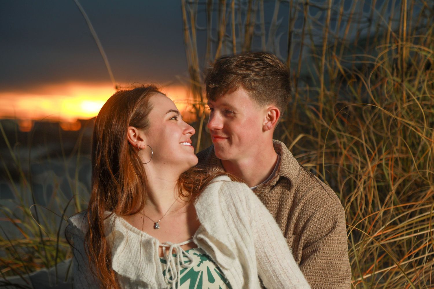 Couple sharing a romantic moment at sunset on a beach, surrounded by golden dune grass.