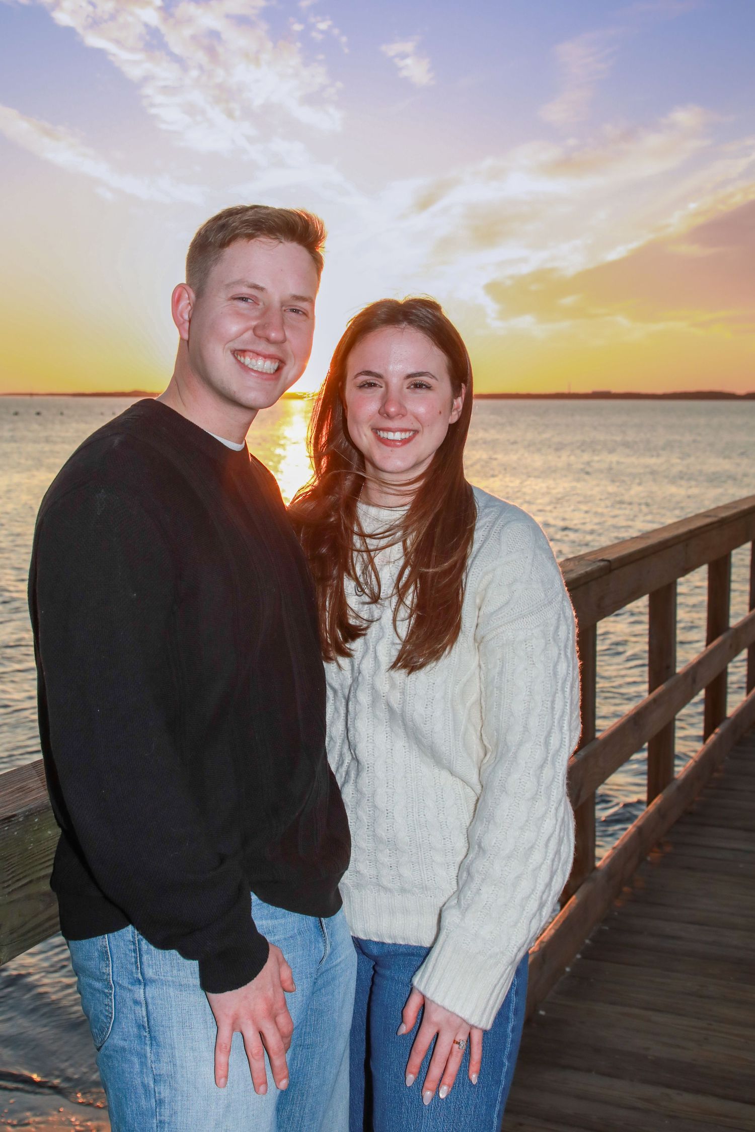 Smiling couple posing on a wooden pier at sunset with golden sky and water in the background.