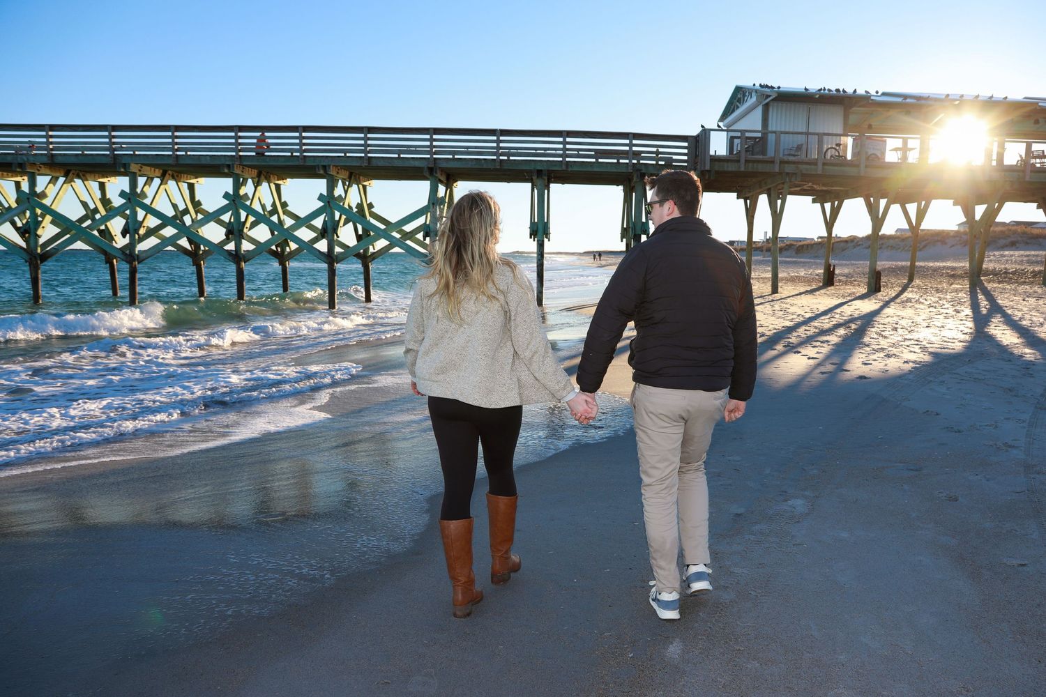 Couple holding hands walking on beach near pier at sunset, woman in brown boots and white sweater.