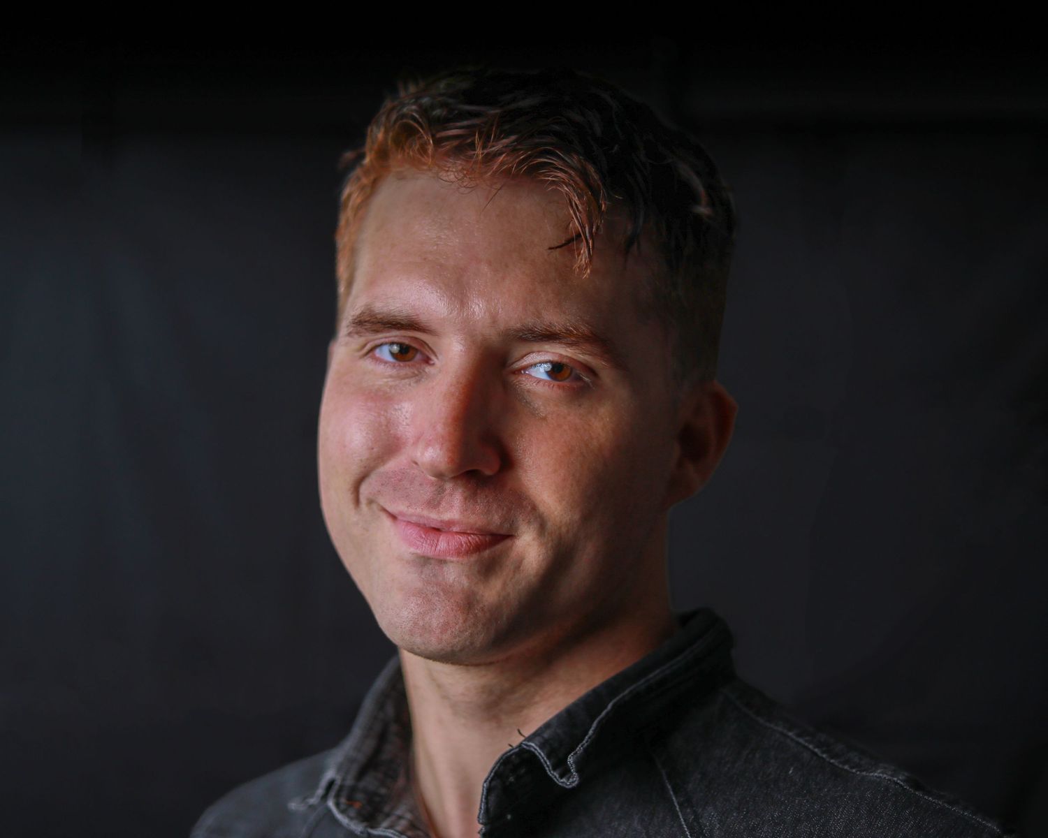 Young man with short reddish hair smiling warmly, wearing a dark gray shirt against a dark background.