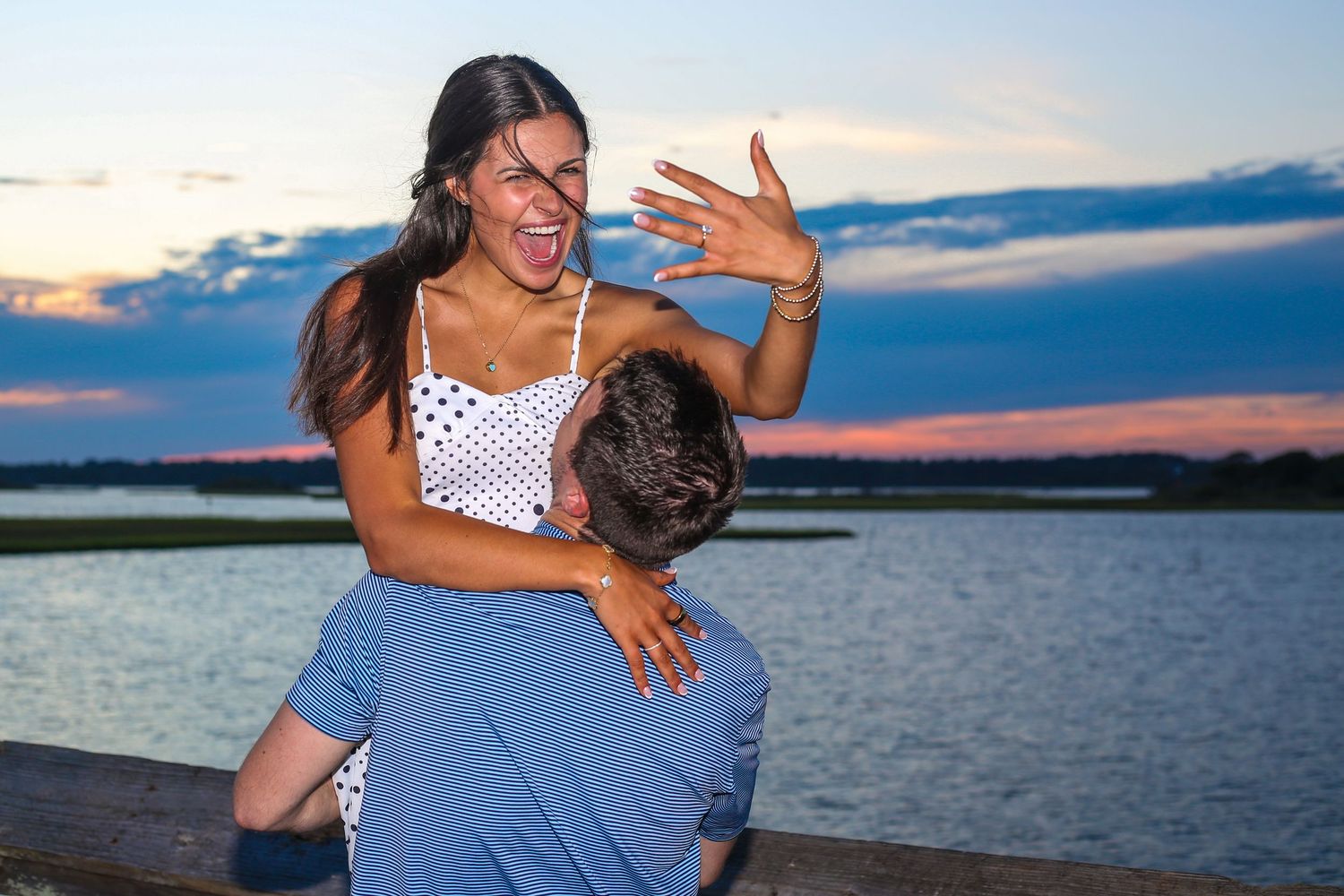 Couple embracing at sunset by a serene lake with colorful sky in background.