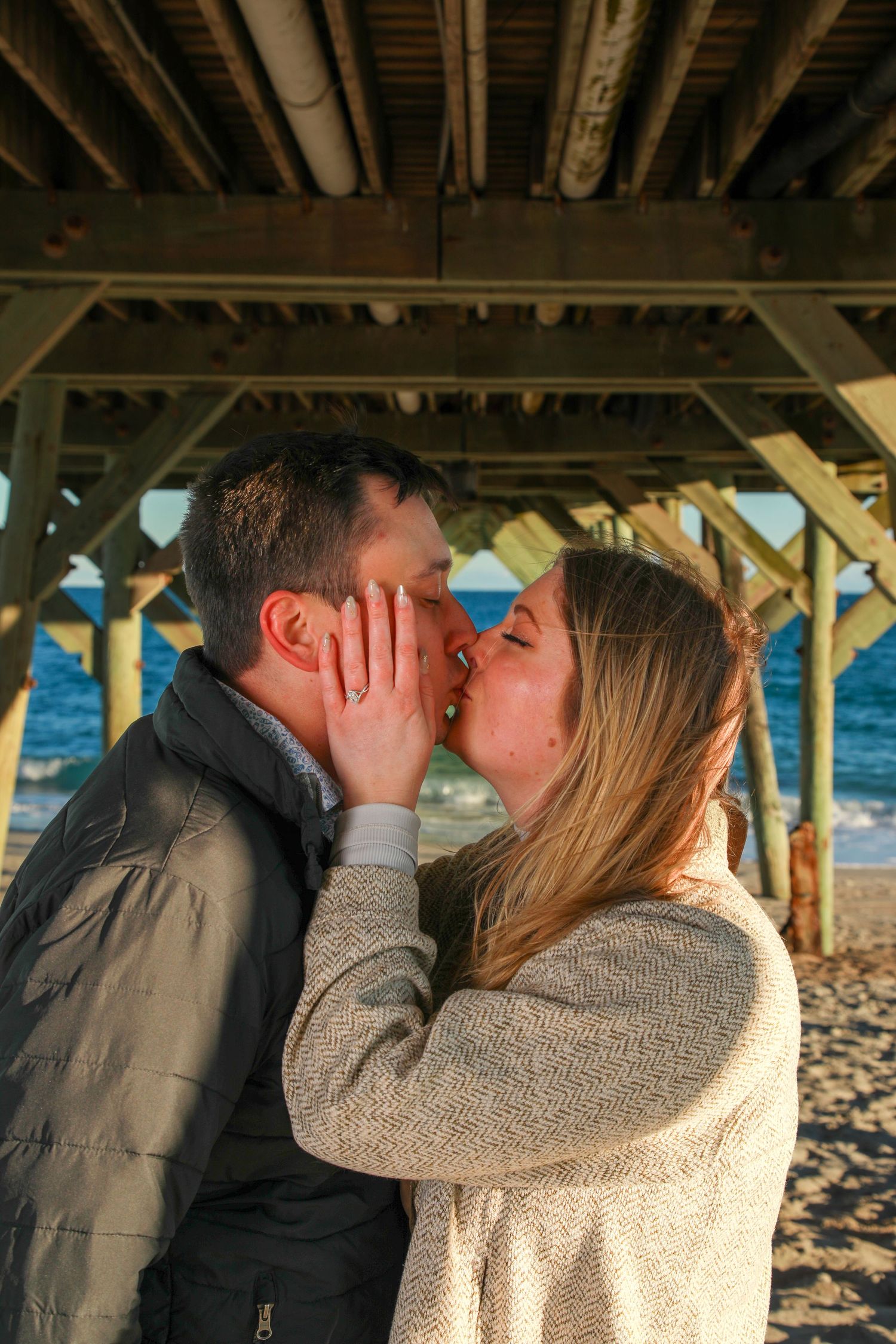 Couple sharing a romantic kiss under a wooden beach pier with ocean waves in the background.