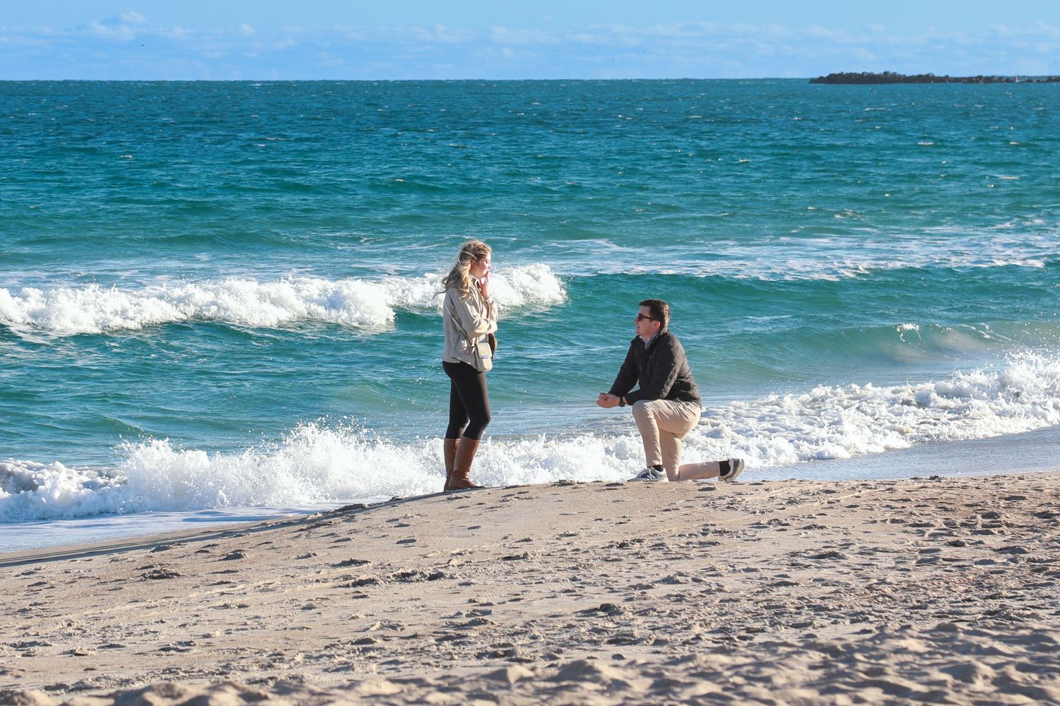 Man proposing on bended knee to woman on sandy beach with turquoise ocean waves in background.