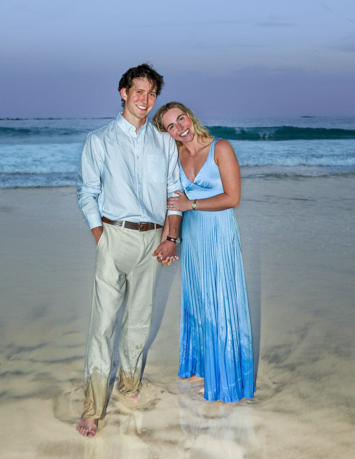 Couple smiling and holding hands on a sandy beach at sunset, man in light blue shirt and woman in blue maxi dress.