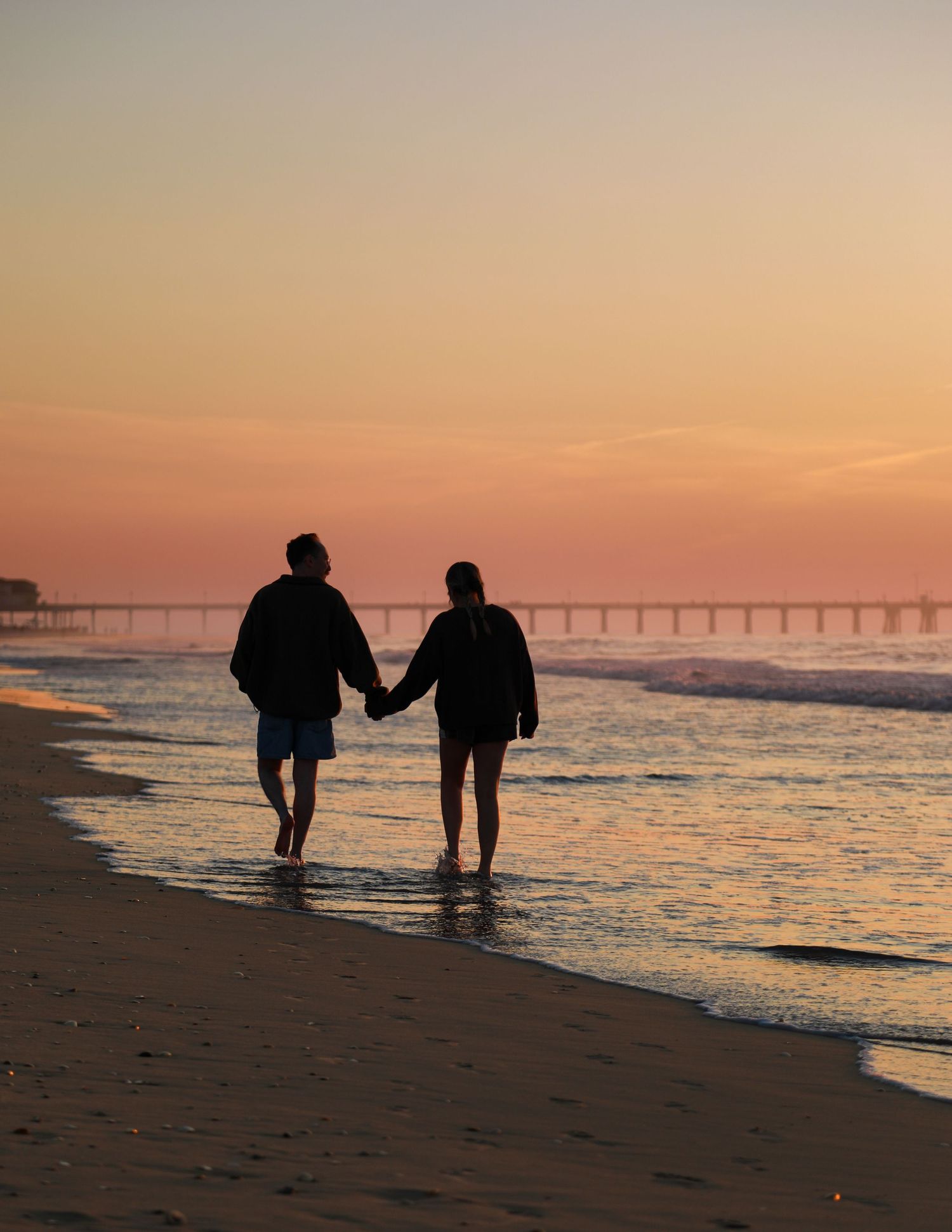 Couple holding hands walking on beach at sunset with pier silhouette in golden orange sky backdrop.