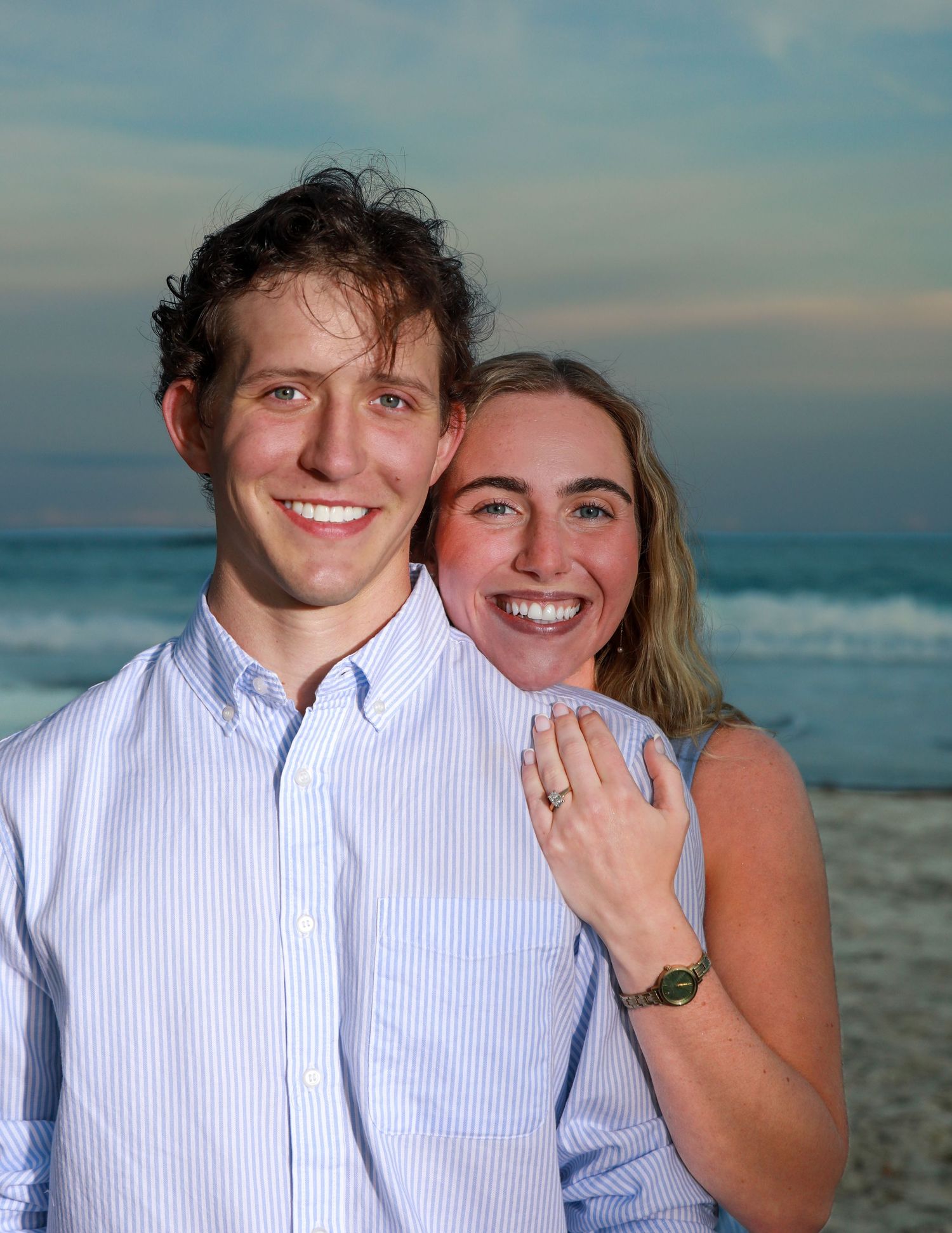 Happy couple smiling at beach during sunset, woman showing engagement ring, wearing casual blue outfits.