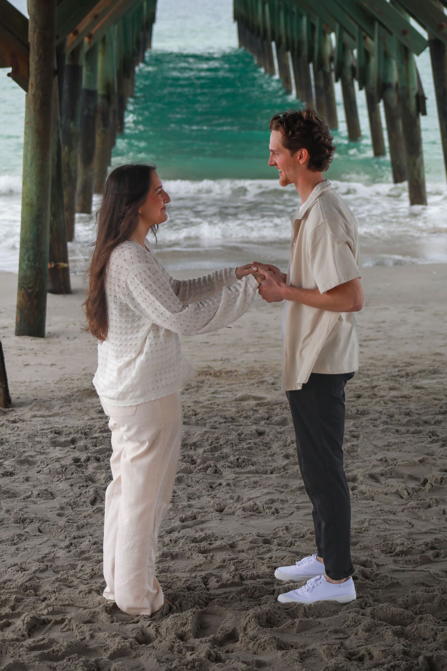 Couple holding hands on sandy beach near pier with ocean waves in background.