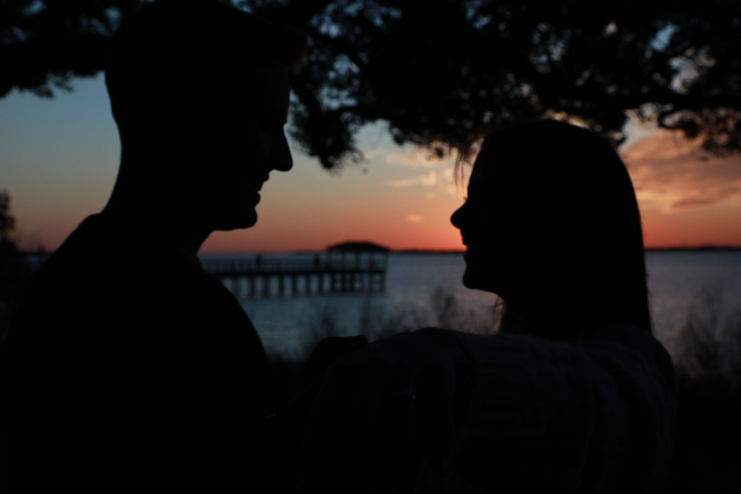 Silhouette of a couple facing each other at sunset with a pier and water in the background.