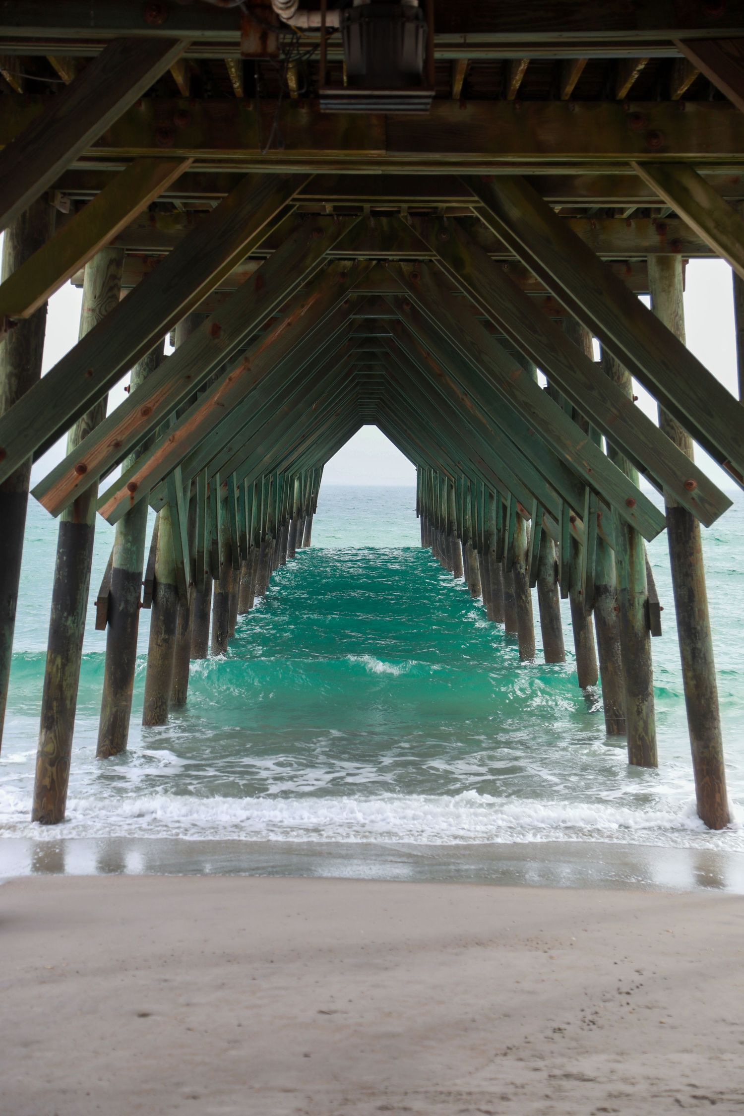 View from beneath a wooden beach pier with turquoise ocean waves and sandy shore, creating a symmetrical tunnel perspective.