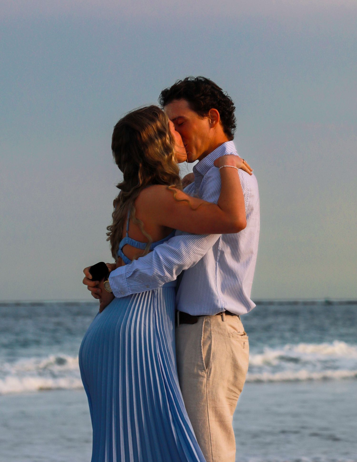 Couple sharing a romantic kiss on the beach at sunset, woman in blue pleated dress, man in white shirt.