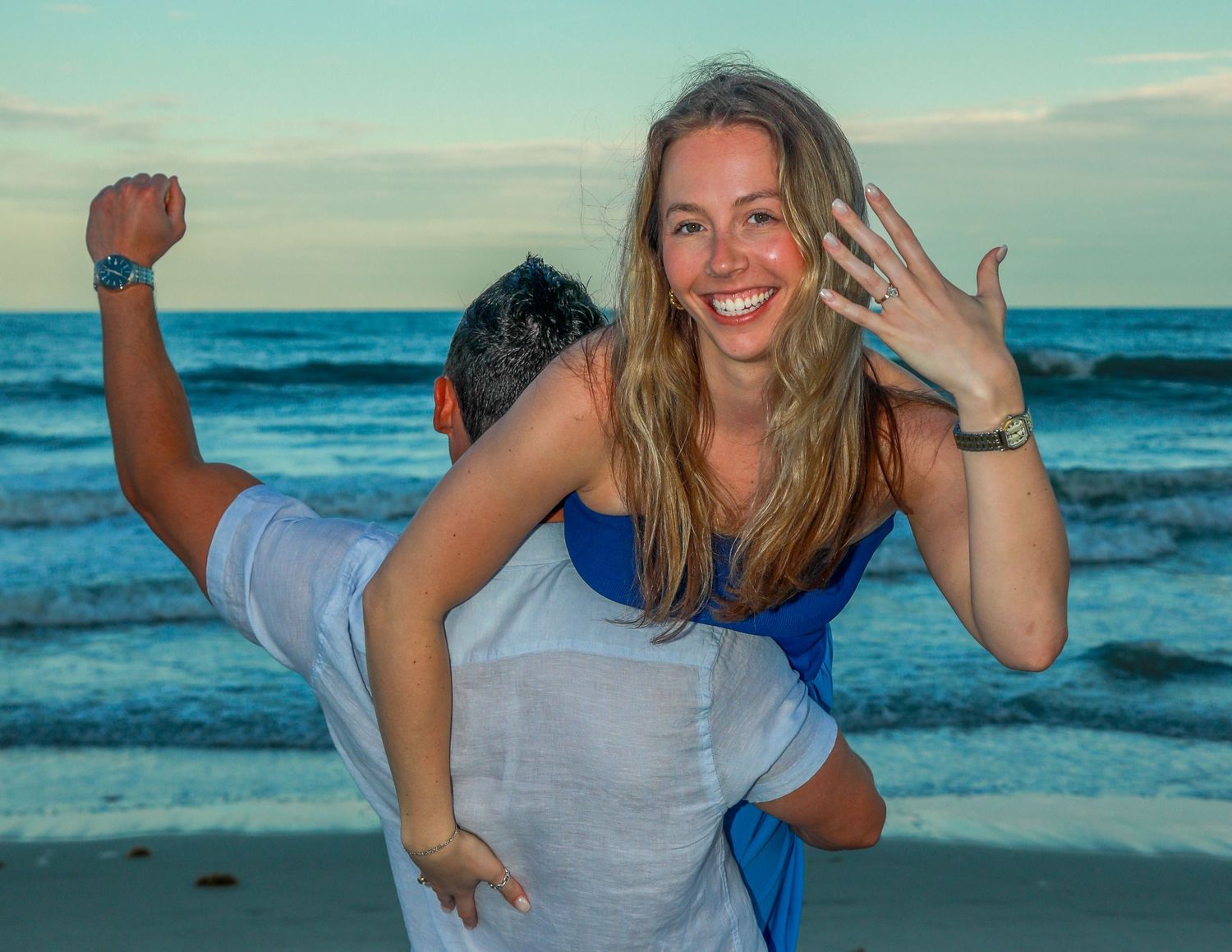 Couple celebrating beach engagement, woman showing ring on piggyback ride at sunset by ocean waves.