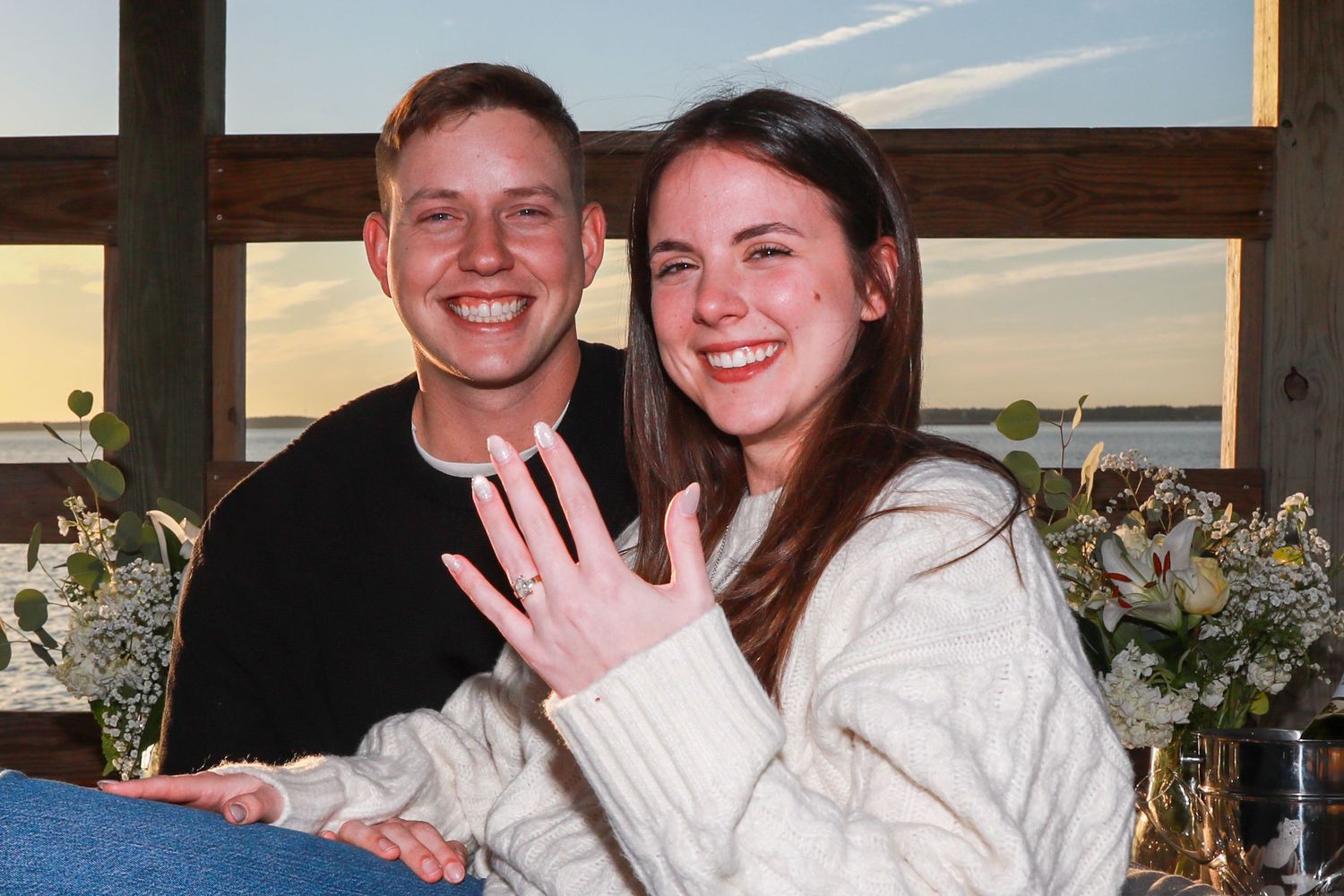 Happy couple smiling at camera showing engagement ring at sunset venue with floral decorations.