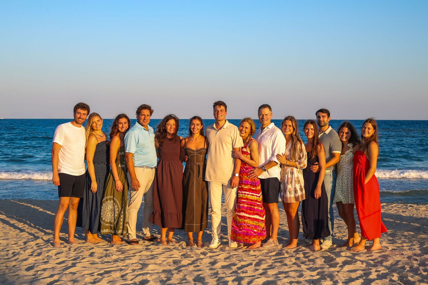 Large group of friends posing together on a sandy beach at sunset with calm ocean waves in the background.