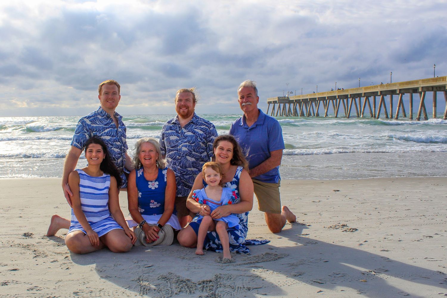Family portrait on a sandy beach near a pier with cloudy skies and ocean waves in the background.