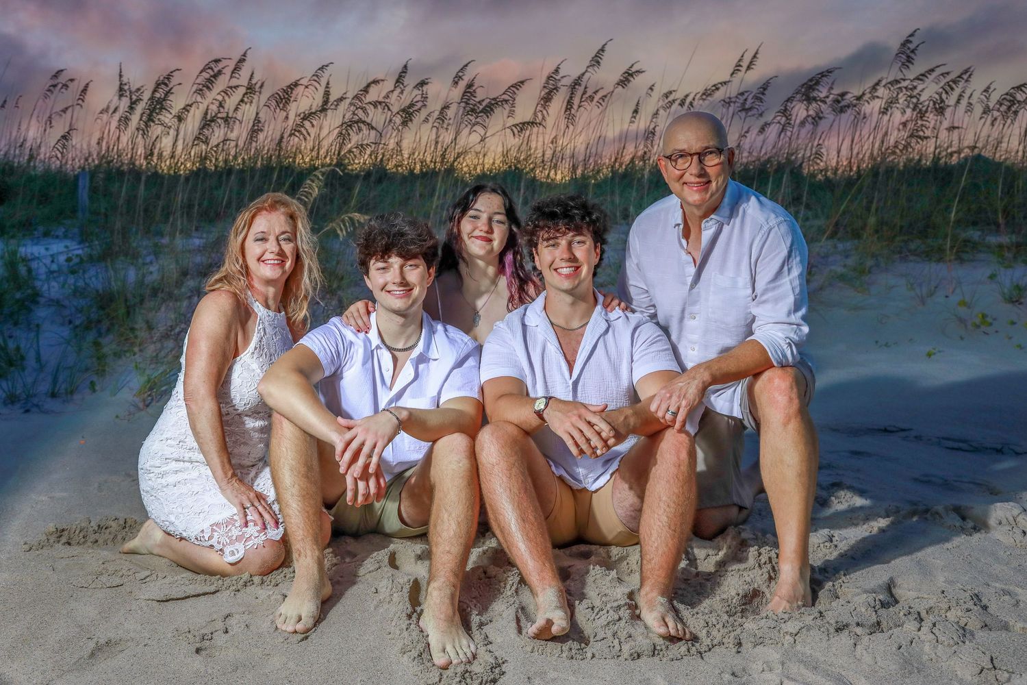 Family of five smiling together on a sandy beach at sunset, wearing white outfits with sea oats in the background.