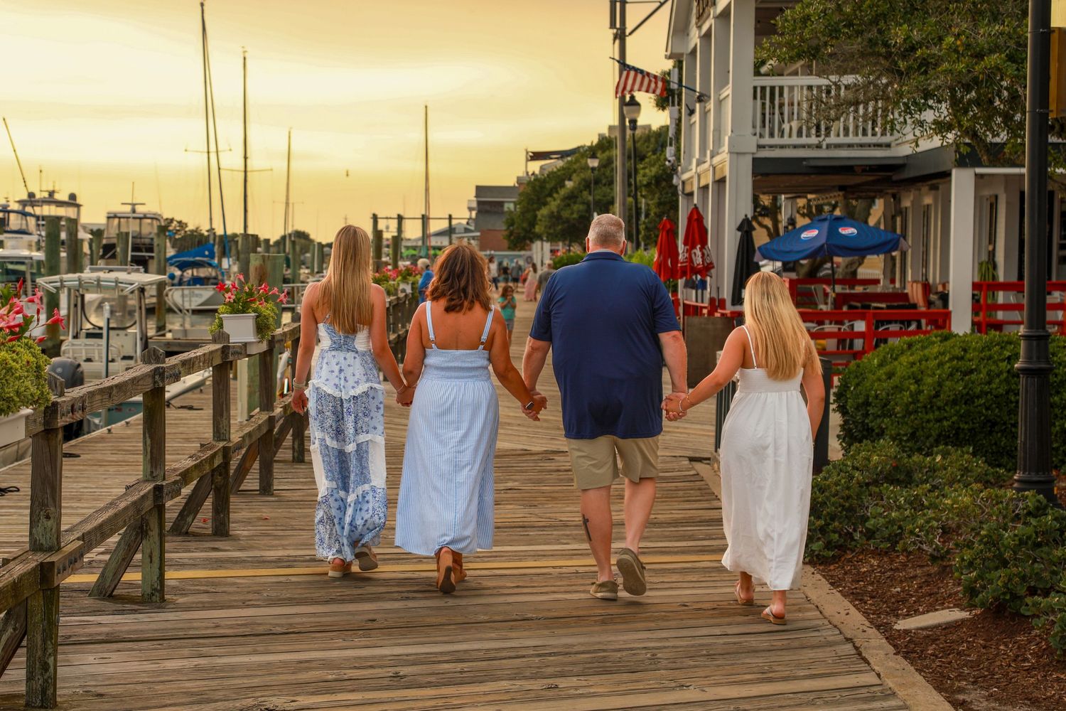 Family of four holding hands walking on a waterfront boardwalk at sunset near a marina.