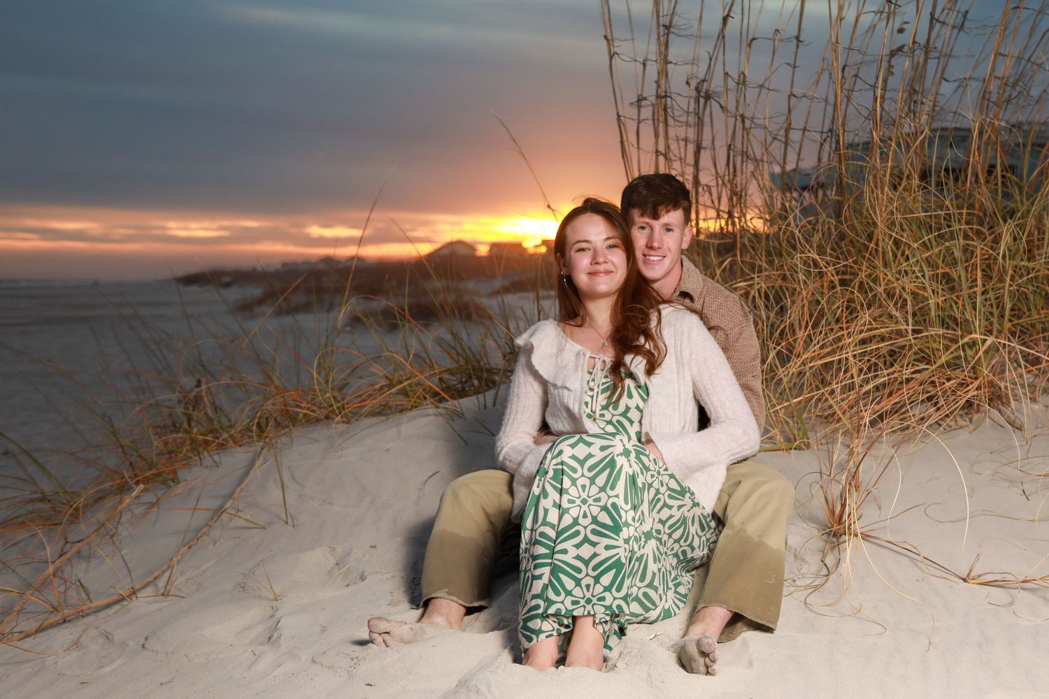 Couple sitting together on sandy beach dunes at sunset, woman in floral dress, man in white sweater, romantic coastal scene.