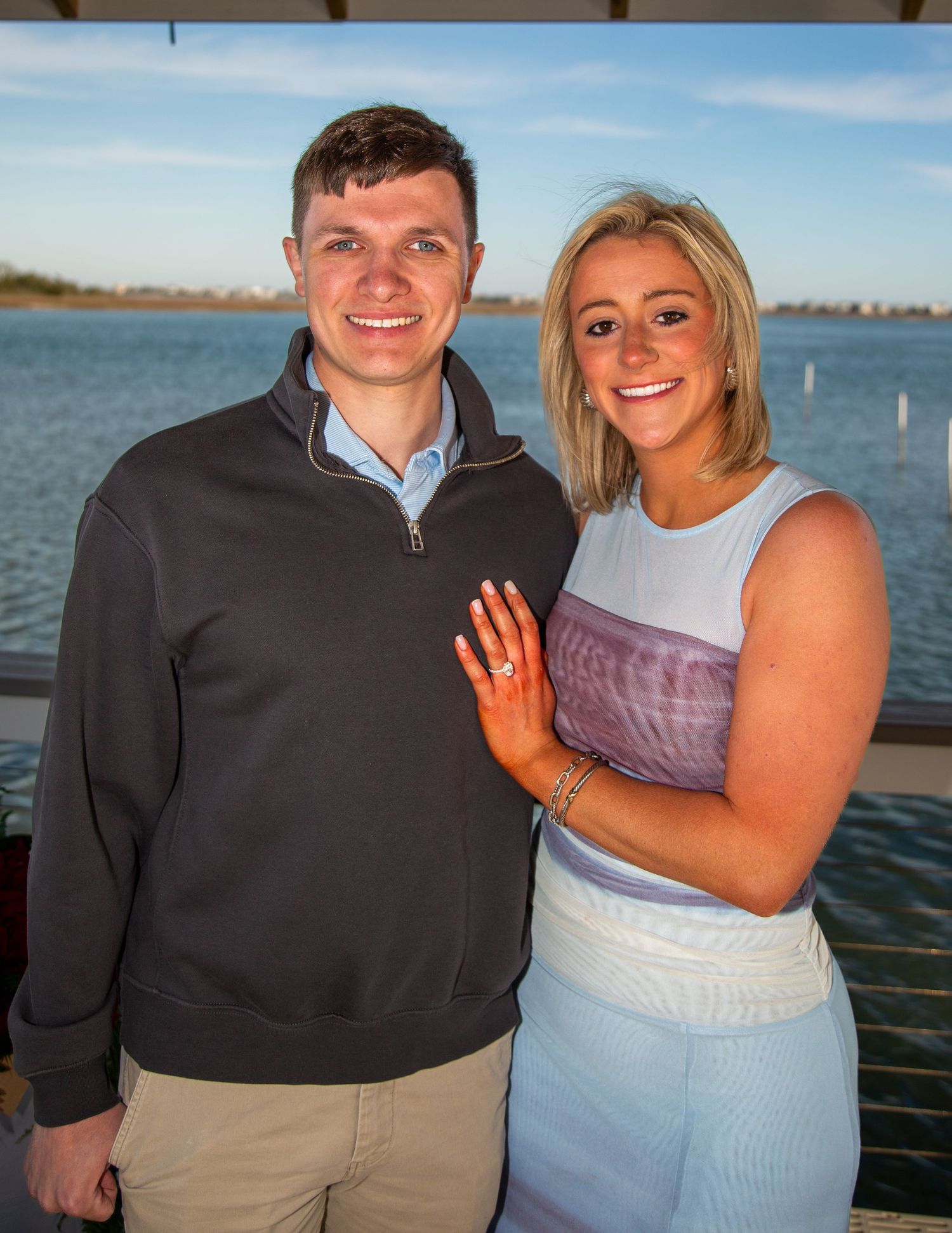 Happy couple posing together outdoors by a waterfront, woman displaying engagement ring, dressed in casual elegant attire.