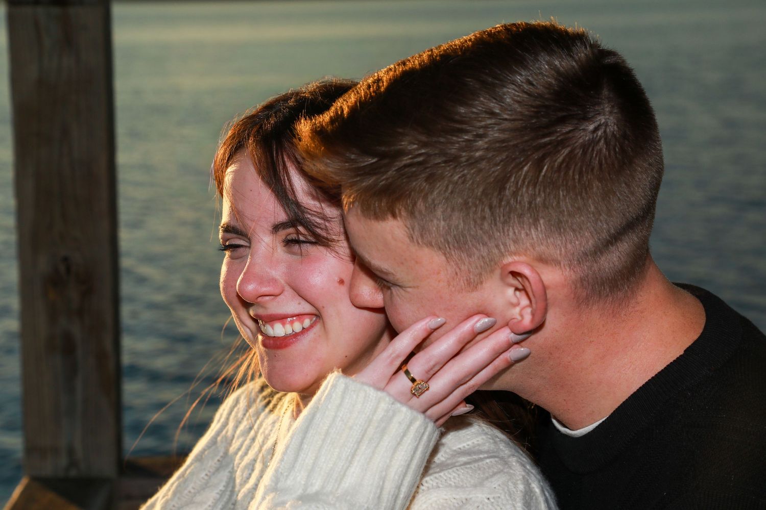 Couple sharing a romantic moment at sunset by the water, woman smiling joyfully wearing a white sweater.