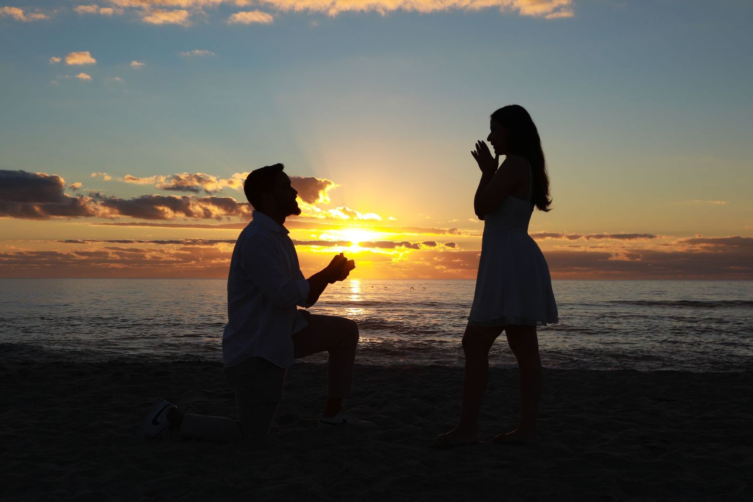 Romantic beach proposal at sunset with man on one knee presenting ring to woman in white dress by the ocean.