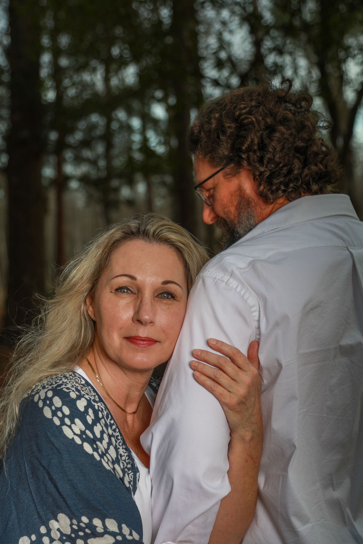 A woman with blonde hair leans against a curly-haired man in glasses outdoors, sharing an intimate moment in a forest.