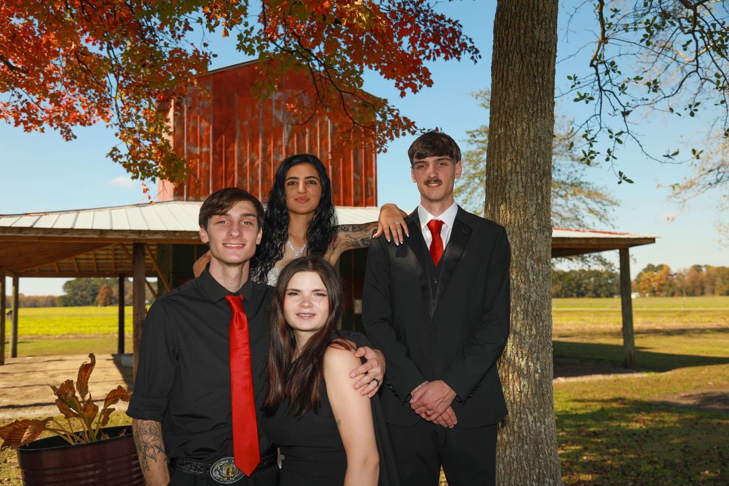 Four teens in formal attire pose outdoors near autumn trees with a red barn in the background.