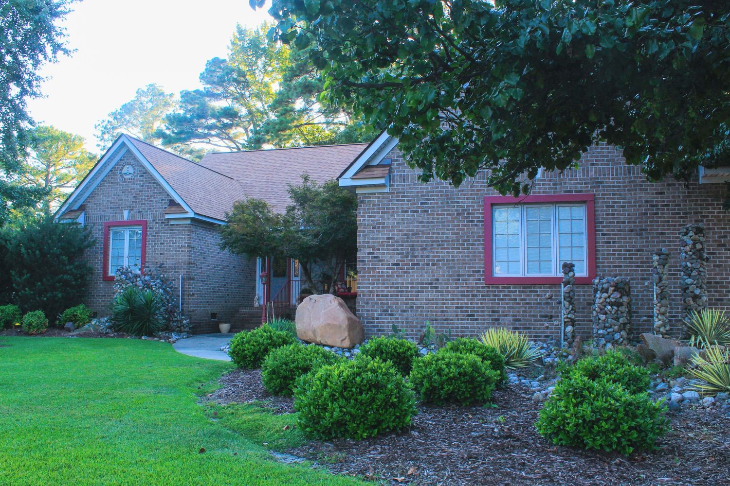 Brick ranch home with red-trimmed windows, lush green lawn, manicured shrubs, and mature trees in a well-landscaped yard.