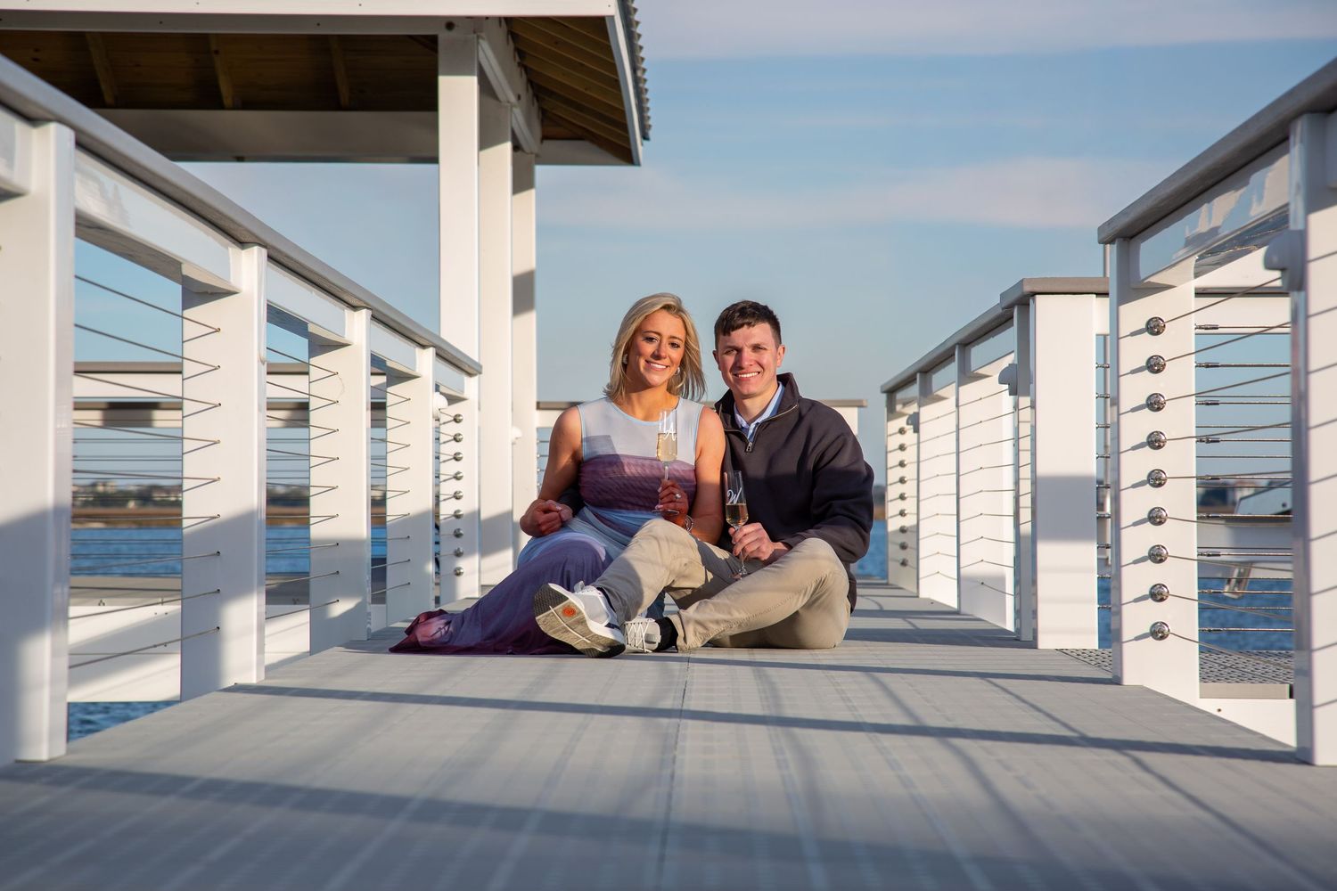 Happy couple sitting together on a white waterfront pier boardwalk under a clear blue sky.