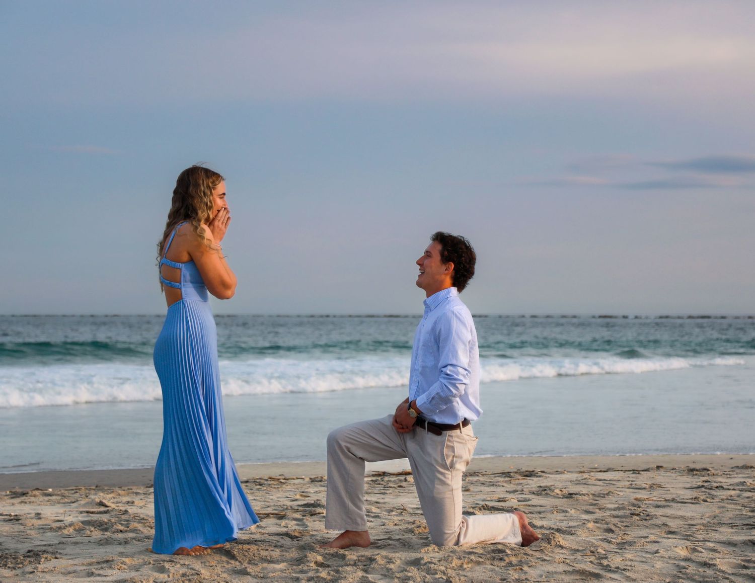 Man proposing on one knee to woman in blue dress on sandy beach at sunset.