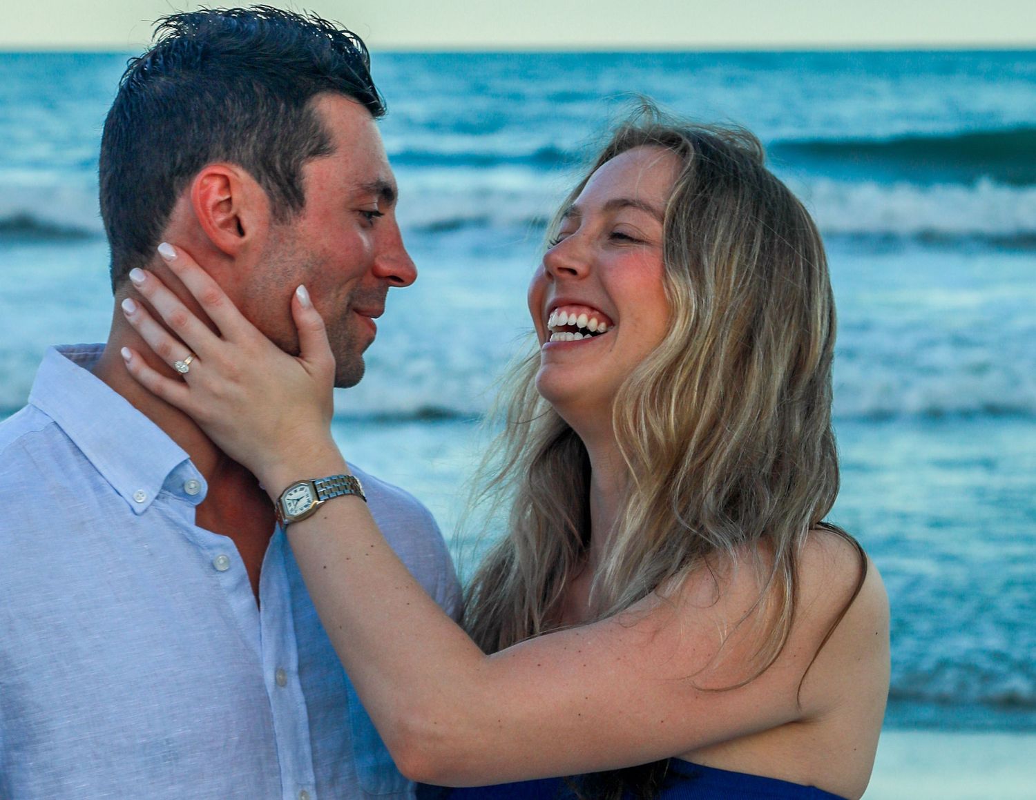 Happy couple embracing and laughing together on a beach at sunset, woman wearing engagement ring.