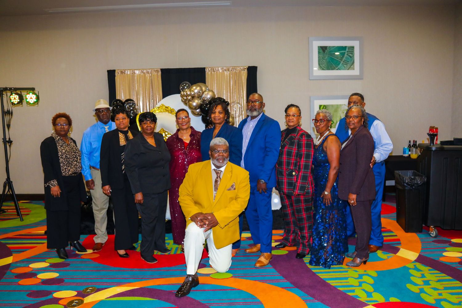 Group of elegantly dressed people posing together at a colorful indoor event venue celebration.