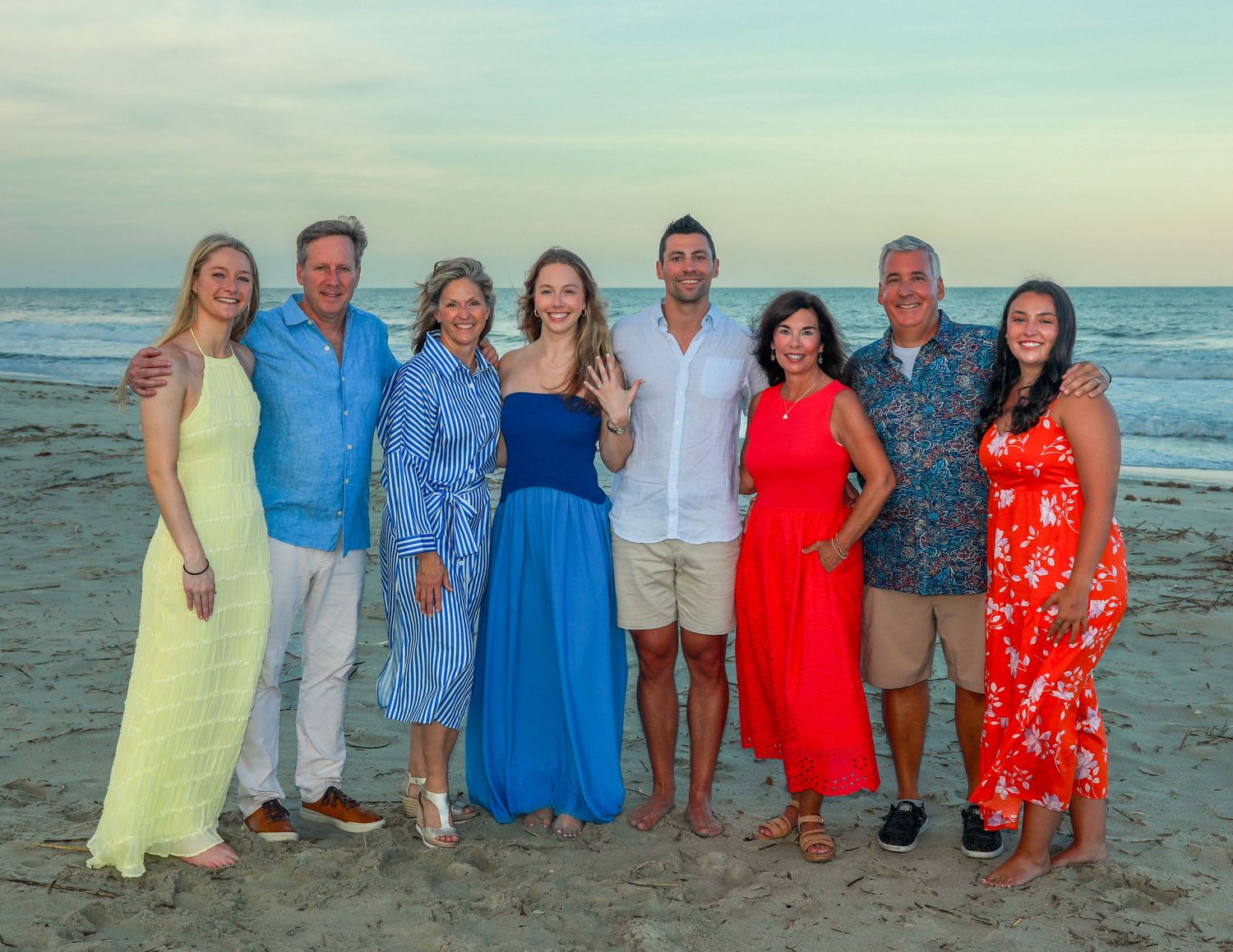 A joyful family group of eight poses together on a sandy beach at sunset, dressed in colorful summer outfits.