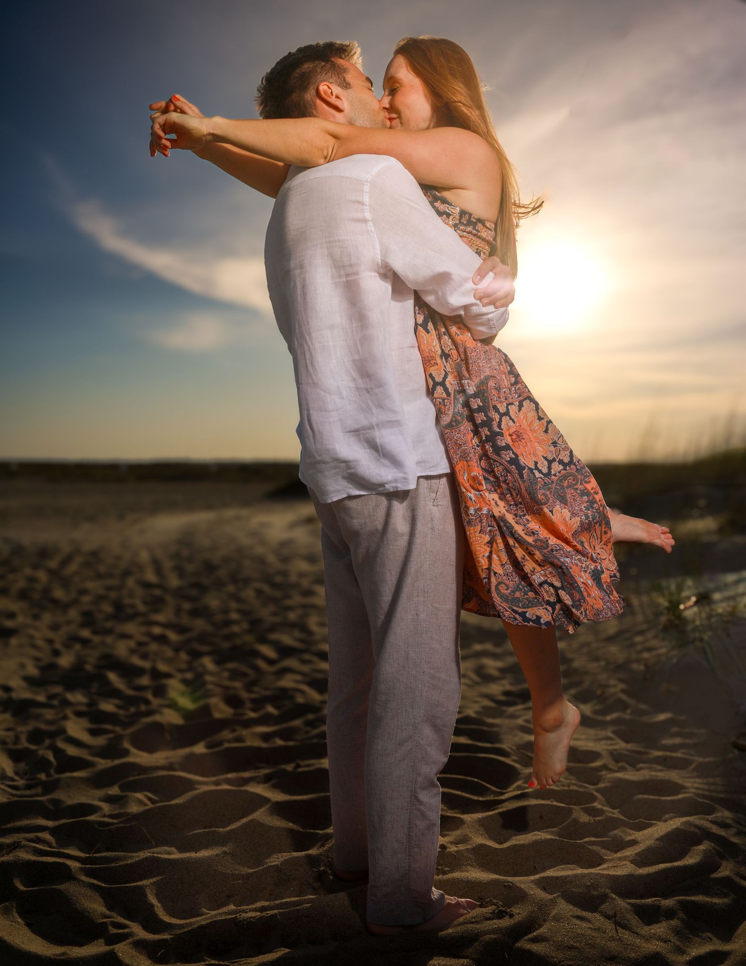 Couple embracing and kissing on a sandy beach at sunset, man in white linen shirt and woman in floral dress.