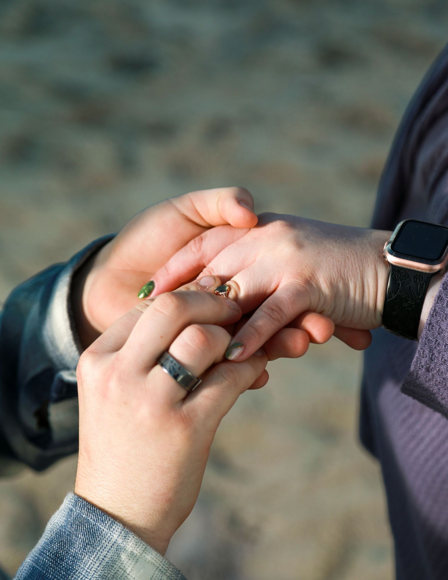 Two people holding hands on a beach, one wearing a black ring and smartwatch, the other with green and gold rings.