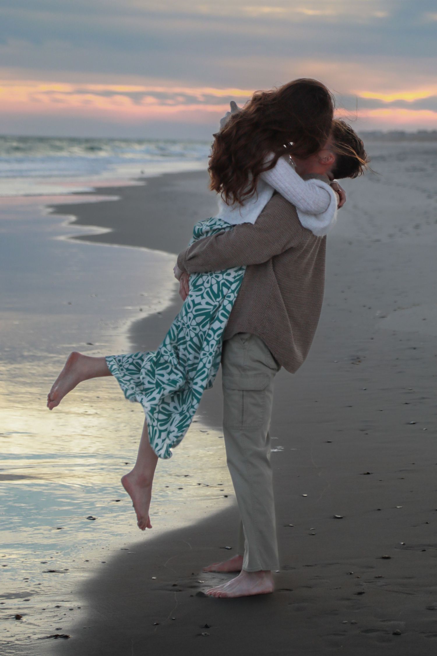 Couple embracing and kissing on a sandy beach at sunset, woman lifted off ground wearing green floral dress.