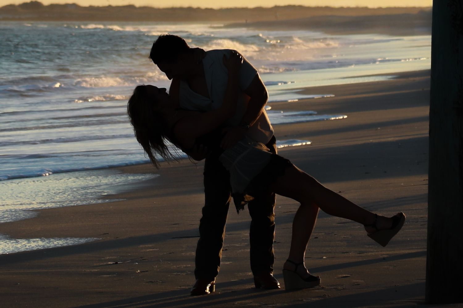 Romantic couple sharing a passionate kiss on a beach at sunset, silhouetted against ocean waves.