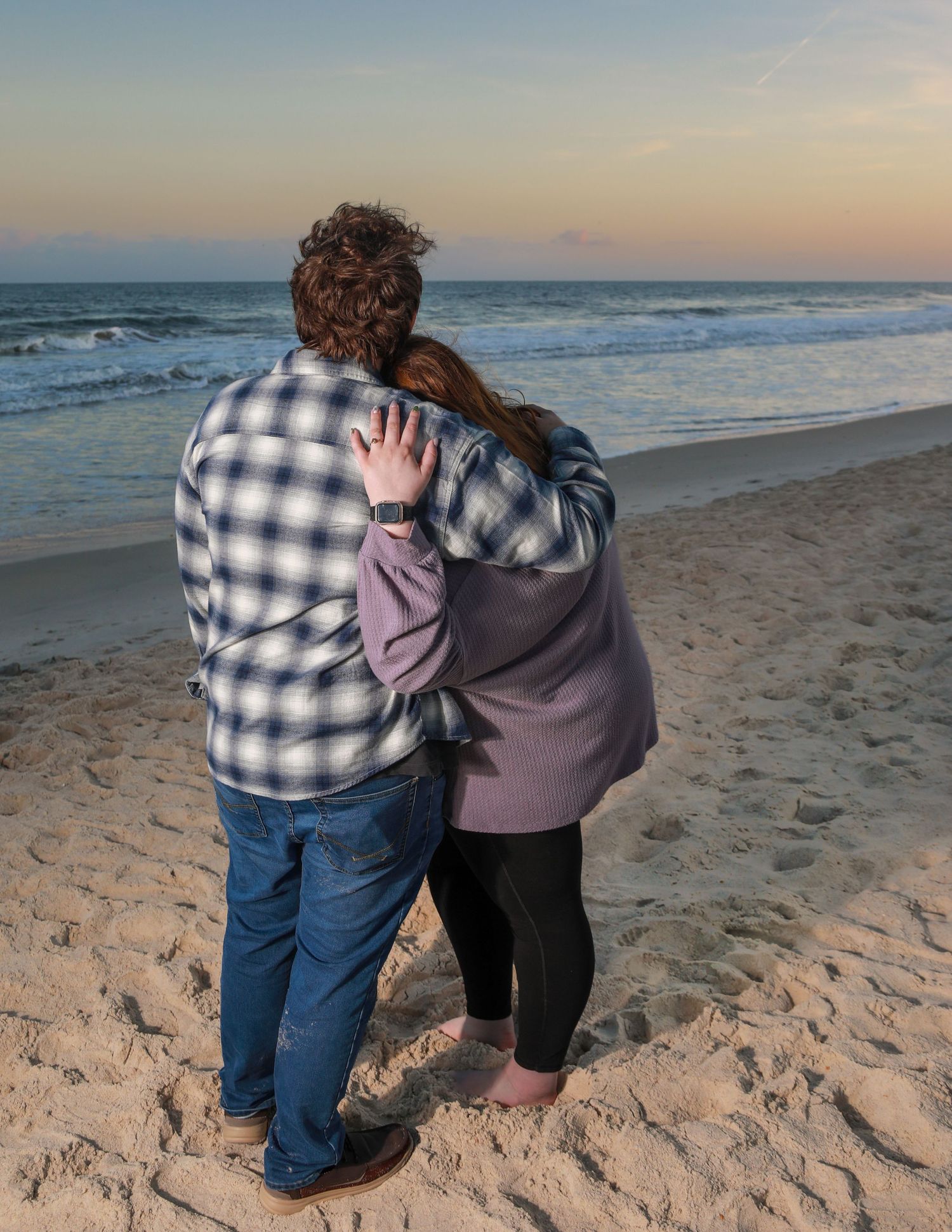 Couple embracing on sandy beach at sunset, watching ocean waves in casual winter clothing.