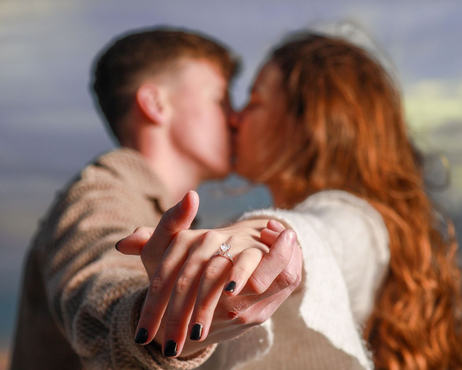 Couple kissing while showing off engagement ring with diamond, woman with red hair outdoors.
