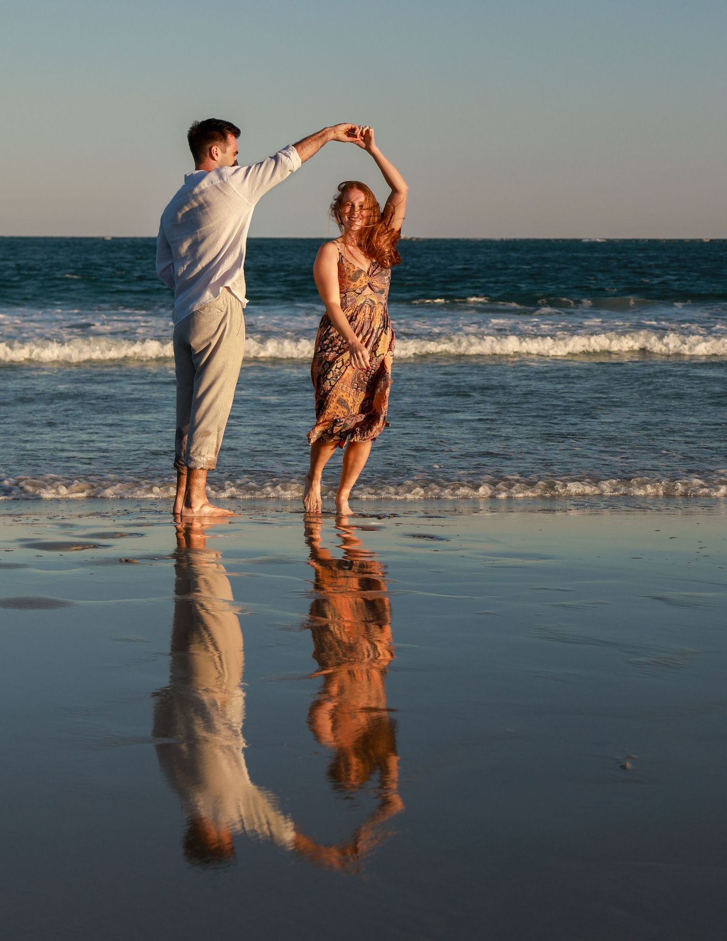 Couple dancing barefoot on a reflective wet beach at sunset, waves in the background.