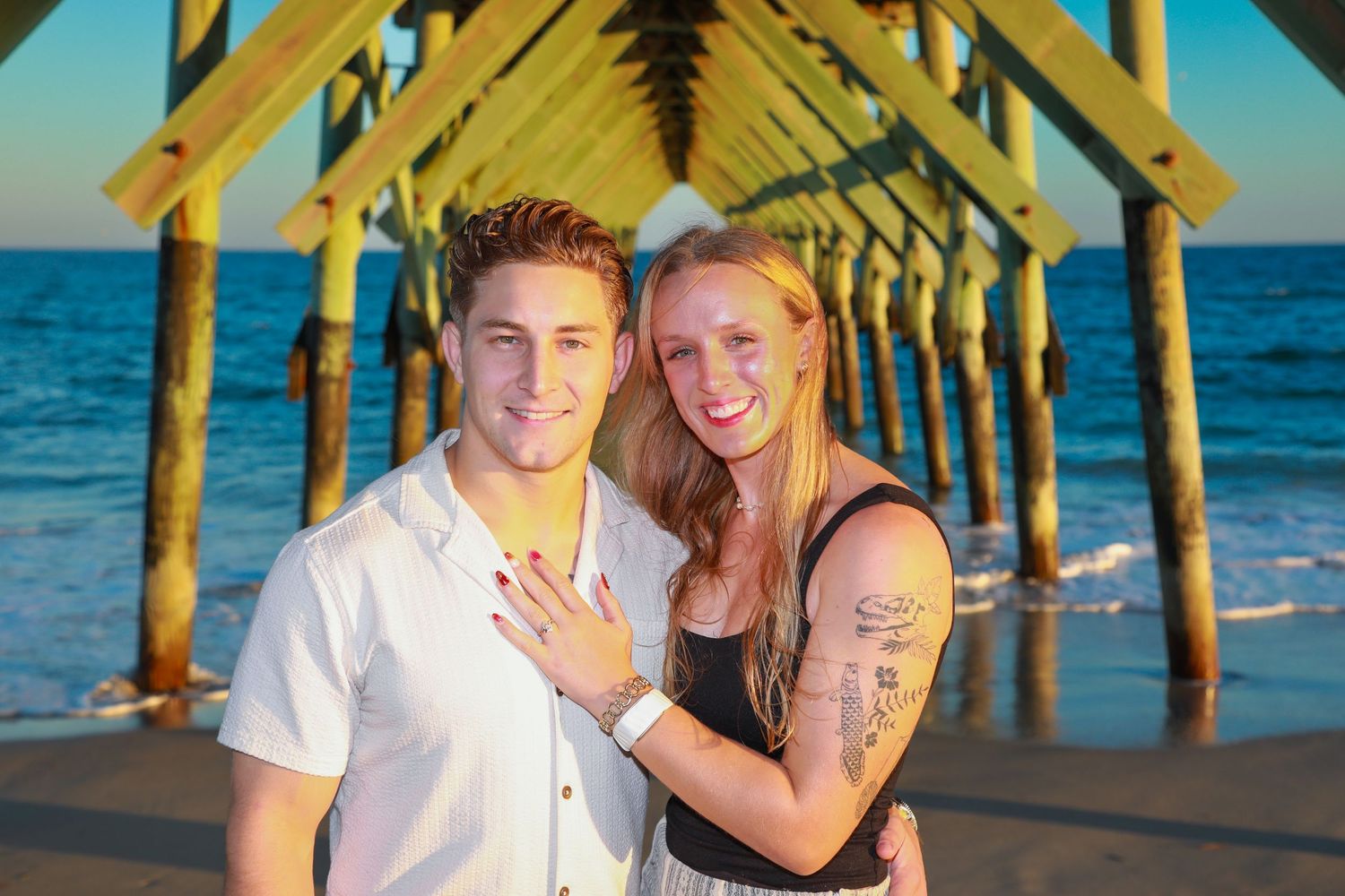 Couple smiling at beach pier, woman showing engagement ring, ocean backdrop at sunset.
