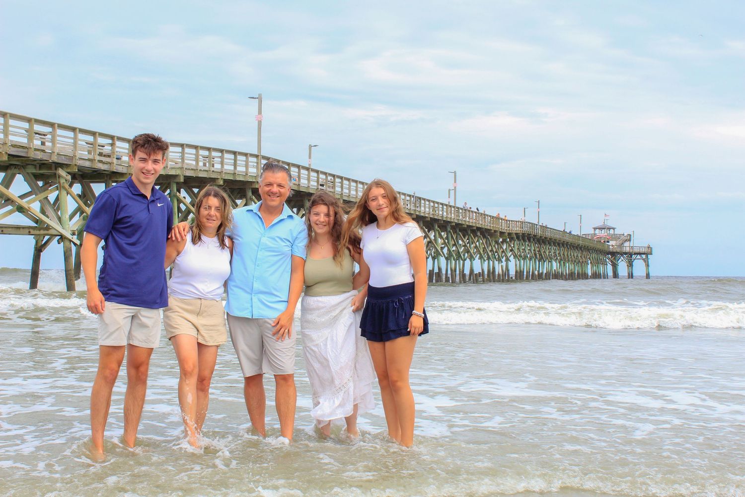 Family of five smiling together on a sandy beach near a pier with ocean waves, on a sunny day.